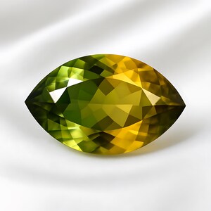 May include: A marquise-cut gemstone, displaying a colour gradient from green to yellow. The gem is faceted, creating a sparkling effect. The background is a soft, white fabric.