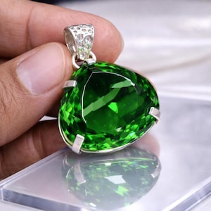 Puede incluir: Colgante de plata con una piedra preciosa verde triangular facetada. La piedra preciosa es de un vibrante color verde esmeralda y está sujeta por garras de plata. La fianza también es de plata y tiene un diseño decorativo.
