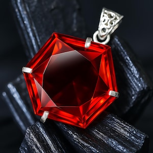 Peut inclure: Pendentif en forme de gemme rouge hexagonale et facettée, avec une bélière en argent. Le pendentif est d'un rouge profond et riche, serti de griffes en argent. La bélière présente un motif ajouré orné. Le pendentif est présenté sur un fond sombre et texturé.