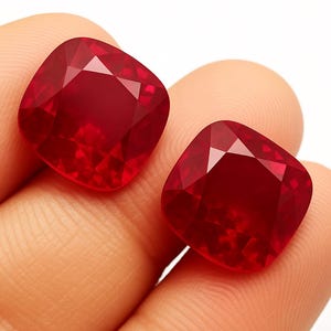 Può includere: Due rubini taglio a cuscino, di un intenso colore rosso. Le gemme sono sfaccettate per massimizzare la riflessione della luce, mostrando la loro brillantezza. I rubini sono tenuti tra le dita.