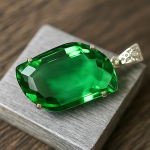 May include: A large, faceted emerald-green gemstone pendant. The pendant has a rectangular shape with a curved edge and is set in a gold-colored metal. The gold setting has a decorative bail. The gemstone is displayed on a gray surface.