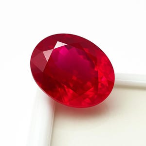 Puede incluir: Una gema de rubí facetada, de forma ovalada. La gema es de un color rojo intenso y vibrante, con múltiples facetas que capturan y reflejan la luz. El rubí está sobre un fondo blanco.