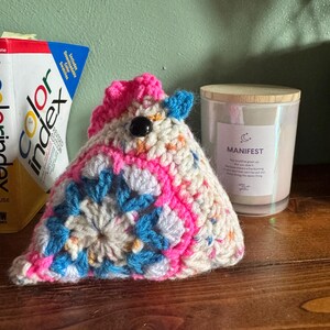 May include: A handmade crochet unicorn with a pink mane and accents of blue and white. The unicorn has a black button eye and is sitting on a wooden surface. A candle with the word "MANIFEST" is in the background.