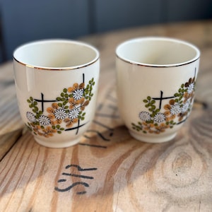 May include: Two off-white ceramic cups with a floral design in orange, white, and green. Each cup has a gold rim and a black cross-like pattern. The cups are on a wooden surface.