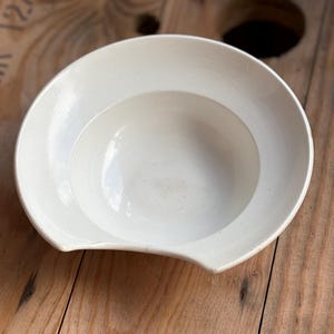 Op de afbeelding: Een witte keramische kom met een brede rand en een gebogen inkeping aan één kant. De kom heeft een ondiepe, afgeronde binnenkant. De kom staat op een houten oppervlak.