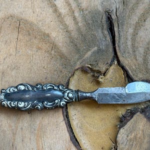 May include: A small, antique silver butter knife with an ornate handle. The handle features a detailed, swirling design. The blade is a flat, triangular shape. The knife is resting on a rough, wooden surface.