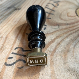 Könnte beinhalten: Ein antiker Wachssiegelstempel mit einem schwarzen Holzgriff und einem Messingstempel. Der Stempel ist mit den Initialen "M.H.2" in einem rechteckigen Rahmen graviert. Der Stempel befindet sich auf einer Holzoberfläche.