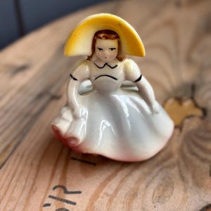 Puede incluir: Figurita de porcelana vintage de una mujer con un sombrero amarillo de ala ancha y un vestido blanco con detalles negros. La figura está pintada a mano y representa a una mujer sentada. La base del vestido es rosa. Un artículo decorativo encantador.