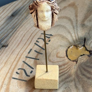 May include: A small, sculpted bust of a woman's face with flowing hair and a blue headpiece. The bust is mounted on a thin, gold-colored rod, which is attached to a small, square, beige base. The sculpture has a weathered, antique appearance.