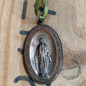 Pode incluir: Pingente religioso oval com uma imagem detalhada de um santo, suspenso por uma fita verde. O pingente apresenta texto em relevo ao redor da borda, e o santo é retratado com os braços estendidos. O pingente é feito de metal cor de bronze.