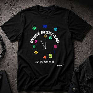 May include: Black t-shirt with a clock design. The clock face has colorful numbers and the words "STUCK IN JET-LAG" in white. Below the clock face, the text reads "-MIND EDITION."