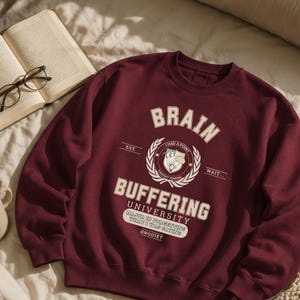 Może przedstawiać: Kasztanowa bluza z okrągłym dekoltem z kremową grafiką z napisem BRAIN BUFFERING UNIVERSITY, EST., WAIT..., I HAD A POINT, THOUGHT PENDING, MAJOR IN FORGETTING WHAT I WAS SAYING oraz &Oslash;NQUIET, wyśrodkowaną wok&oacute;ł emblematu wieńca laurowego.