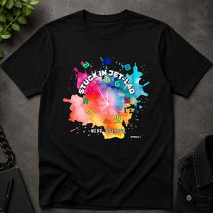 May include: Black t-shirt featuring a colorful watercolor design with the words "STUCK IN JET-LAG" and a clock face. The design includes the text "-MIND EDITION" and a small signature.