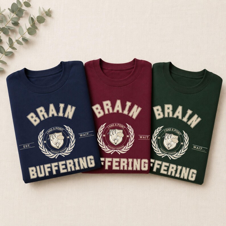Może przedstawiać: Trzy bluzy z okrągłym dekoltem w kolorze granatowym, bordowym i ciemnozielonym. Każda posiada kremową grafikę z napisem "BRAIN BUFFERING", centralnym herbem z ikoną żar&oacute;wki oraz frazą "I HAD A POINT THOUGHT PENDING."