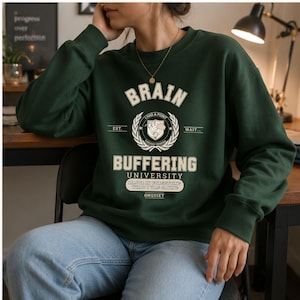 Może przedstawiać: Ciemnozielona bluza z okrągłym dekoltem i kremowym nadrukiem z napisem BRAIN BUFFERING UNIVERSITY, I HAD A POINT, WAIT, EST, THOUGHT PENDING, MAJOR IN FORGETTING WHAT I WAS SAYING oraz &Oslash;NQUIET, umieszczonym centralnie na klatce piersiowej.