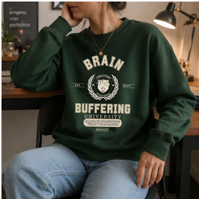 Może przedstawiać: Ciemnozielona bluza z okrągłym dekoltem i kremowym nadrukiem z napisem BRAIN BUFFERING UNIVERSITY, I HAD A POINT, WAIT, EST, THOUGHT PENDING, MAJOR IN FORGETTING WHAT I WAS SAYING oraz &Oslash;NQUIET, umieszczonym centralnie na klatce piersiowej.