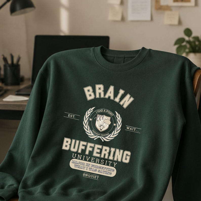 Może przedstawiać: Ciemnozielona bluza z okrągłym dekoltem i kremowym nadrukiem z tekstem BRAIN BUFFERING UNIVERSITY, I HAD A POINT, THOUGHT PENDING, WAIT, MAJOR IN FORGETTING WHAT I WAS SAYING oraz ONQUIET, wyśrodkowanym z tarczą i motywem wieńca laurowego.