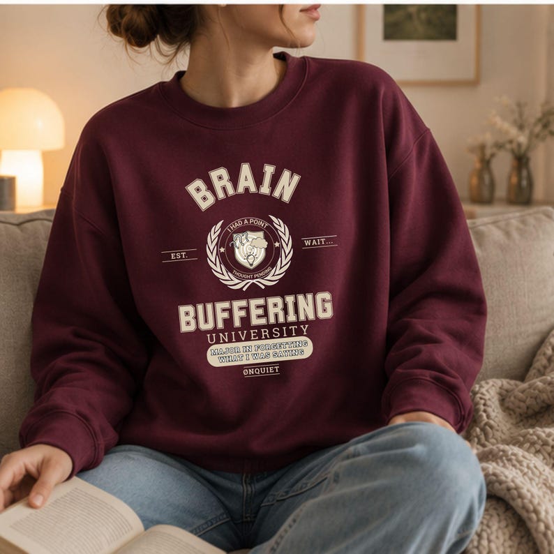 Może przedstawiać: Bordowa bluza z okrągłym dekoltem z kremowym nadrukiem zawierającym tekst BRAIN BUFFERING UNIVERSITY, I HAD A POINT, WAIT, EST, THOUGHT PENDING, MAJOR IN FORGETTING WHAT I WAS SAYING oraz &Oslash;NQUIET, umieszczonym centralnie na klatce piersiowej.