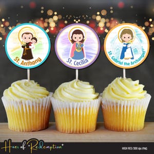 Op de afbeelding: Drie cupcakes met decoratieve toppers met illustraties van St. Antonius, St. Cecilia en St. Gabriël de Aartsengel. De toppers zijn rond met kleurrijke ontwerpen en tekst. De cupcakes hebben gele frosting.