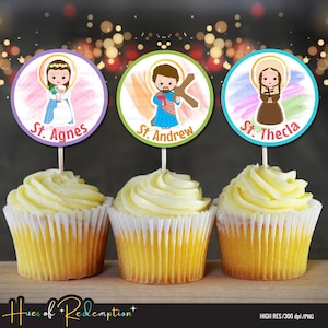 Op de afbeelding: Drie cupcakes met gele glazuur, elk bekroond met een rond beeld van een heilige. De afbeeldingen zijn gelabeld met "St. Agnes", "St. Andrew" en "St. Thecla". De cupcakes staan op een houten oppervlak.