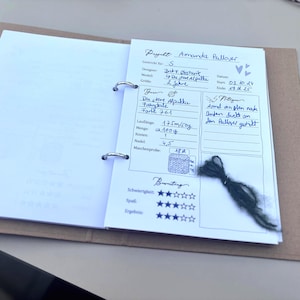 Könnte beinhalten: Offenes Notizbuch mit handschriftlichen Notizen zu einem Strickprojekt, dem "Amanda Pullover". Die Seite enthält Angaben zu Garn, Farbe und Nadelstärke. Ein kleines Strickmuster und ein Bündel grünes Garn sind ebenfalls zu sehen.