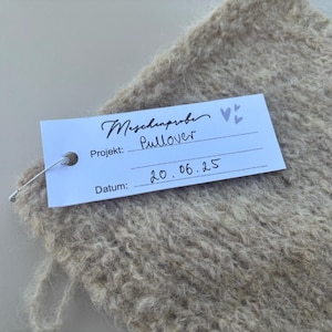 May include: Close-up of a beige knitted fabric with a tag attached. The tag reads "Maschenprobe" and "Pullover" with the date "20.06.25". The fabric has a textured, fluffy appearance, suggesting a soft material.