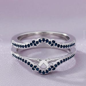 May include: A silver ring with a double band design. The bands are adorned with small, dark blue gemstones. The ring has a curved shape, with the gemstones following the curve. The background is a soft, light purple fabric.