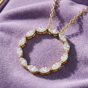 Puede incluir: Un collar de oro con un colgante circular engastado con piedras transparentes de talla marquesa. El colgante cuelga de una delicada cadena de oro y descansa dentro de una caja de madera oscura forrada con terciopelo morado claro.
