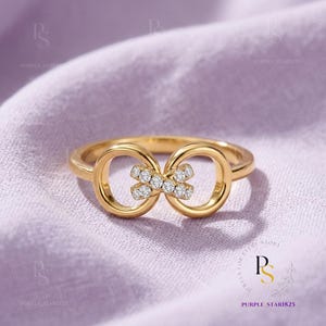 Peut inclure: Une bague dorée avec un motif de symbole infini. La bague présente deux boucles circulaires reliées par un groupe de petits pierres précieuses transparentes. La bague est posée sur un tissu violet clair.