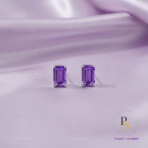 May include: A pair of stud earrings with rectangular, deep purple gemstones set in silver. The earrings are displayed on a soft, lavender-colored fabric, creating a luxurious and elegant presentation. The gemstones have a vibrant, saturated color.