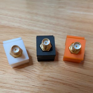 May include: Three small electronic connectors in white, black, and orange. Each has a gold-colored central component and a hexagonal base. The connectors are arranged on a light brown wooden surface.
