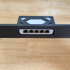 May include: A black network rack mount with a white device featuring multiple Ethernet ports. The rack mount has a rectangular design with mounting holes on each end. The device has several ports and indicator lights.