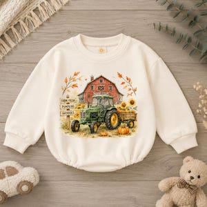 May include: A cream-coloured long-sleeve sweatshirt featuring a graphic print of a red barn, a green tractor, sunflowers, and pumpkins. A wooden signpost displays the text Farm Fresh, Pumpkins, and Hayrides. The garment has a crew neckline.