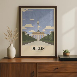 May include: A framed vertical wall art print featuring a stylized illustration of the Berlin TV Tower and the Brandenburg Gate under a blue sky with clouds. The text BERLIN GERMANY is printed in dark blue at the bottom of the cream-colored poster.