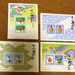 May include: Four rectangular Japanese New Year postage stamp souvenir sheets featuring colourful illustrations of traditional motifs, including a dragon, origami crane, and folk figurines, with Japanese calligraphy and specific era dates.