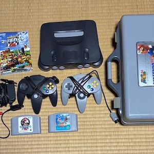 Może przedstawiać: Kompletny zestaw do gier Nintendo 64, zawierający konsolę, kontrolery, kartridże do gier i walizkę. Konsola i kontrolery są czarne i szare. Obudowa jest szara z napisem "NINTENDO 64" z boku.