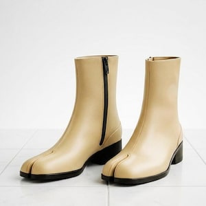 May include: A pair of beige leather ankle boots with a split-toe design. The boots have a black zip and a small black heel. The boots are displayed on a white tiled surface.