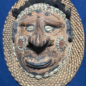 Op de afbeelding: Een handgemaakt, ovaalvormig masker met een lachend gezicht. Het masker is bruin met zwarte accenten en versierd met kleine witte schelpen. Het is geplaatst tegen een geweven, lichtbruine achtergrond met zwarte accenten.