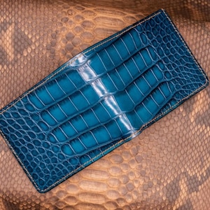 May include: A teal blue leather wallet with a crocodile skin pattern. The wallet is rectangular and lies on a snakeskin-textured surface. The wallet has a stitched border and a glossy finish.