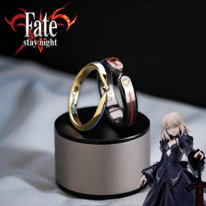 Puede incluir: Dos anillos, uno dorado y otro negro, exhibidos en un soporte plateado. El anillo dorado tiene un detalle de diamante, mientras que el anillo negro presenta detalles rojos y amarillos. El texto "Fate stay night" está presente.