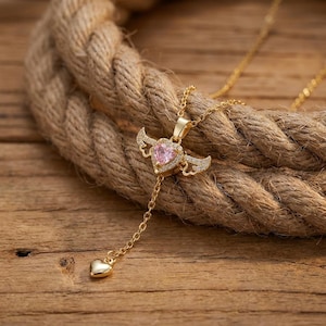 Puede incluir: Un collar de tono dorado con un colgante central en forma de coraz&oacute;n con una piedra rosa y alas decorativas adornadas con peque&ntilde;os cristales transparentes. Una delicada cadena dorada desciende del coraz&oacute;n alado hasta un peque&ntilde;o dije de coraz&oacute;n.