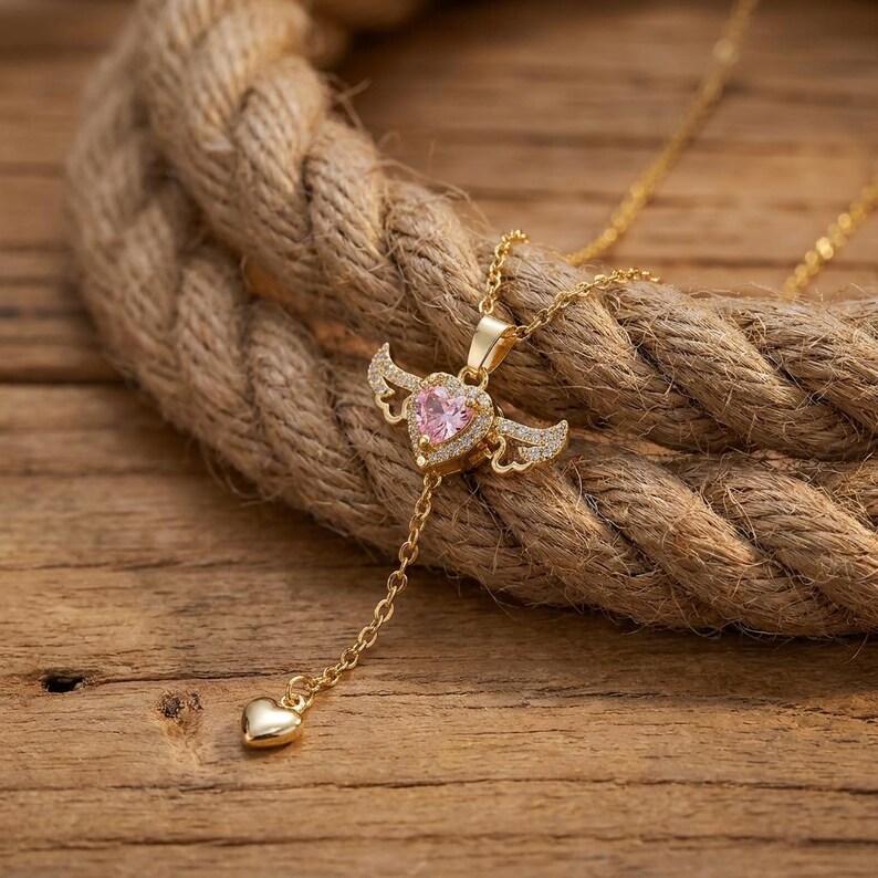 Puede incluir: Un collar de tono dorado con un colgante central en forma de coraz&oacute;n con una piedra rosa y alas decorativas adornadas con peque&ntilde;os cristales transparentes. Una delicada cadena dorada desciende del coraz&oacute;n alado hasta un peque&ntilde;o dije de coraz&oacute;n.