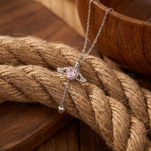 Puede incluir: Un collar de tono plateado con un colgante central de piedra rosa en forma de coraz&oacute;n, adornado con peque&ntilde;as alas y piedras transparentes. Una cadena delicada se extiende hacia abajo desde el coraz&oacute;n, terminando en un peque&ntilde;o dije liso.