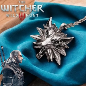 May include: Silver-colored pendant necklace featuring the Witcher's wolf medallion. The pendant is a detailed, stylized wolf head with sharp features. The necklace is displayed on a blue fabric, with the game title "The Witcher: Wild Hunt" visible.