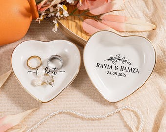 Könnte beinhalten: Zwei herzförmige, weiße Keramik-Ringschalen mit goldenem Rand. Eine Schale enthält einen goldenen Ring, einen silbernen Ring und Ohrringe. Auf der anderen Schale stehen die Namen "Rania & Hamza" und das Datum "24.08.2025".