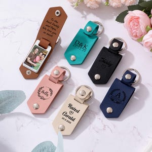 Puede incluir: Una colección de llaveros personalizados de cuero en varios colores, incluyendo marrón, turquesa, negro, rosa, beige y azul marino. Algunos llaveros presentan nombres, iniciales, fechas y una foto grabados. Un llavero tiene el texto: "To the best Daddy and Papa, We love you so much."