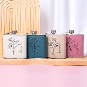 May include: Four stainless steel hip flasks with floral designs and names. The flasks are in white, teal, beige, and pink. Each flask has a different floral illustration and name in black script. The flasks are displayed on a wooden surface.