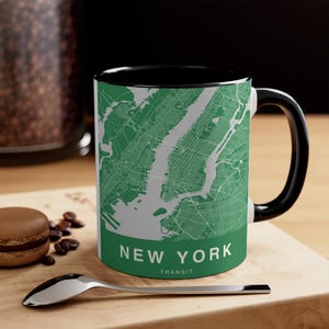 May include: A ceramic mug with a black handle and interior. The exterior features a green map of New York City with the words "NEW YORK TRANSIT" in white. A spoon and a macaron are in the foreground.