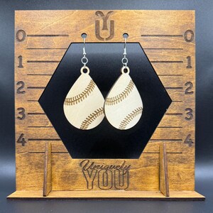 Könnte beinhalten: Baseball-Themen-Ohrringe in Tropfenform. Diese Ohrringe sind aus hellem Holz gefertigt und zeigen ein Baseball-Design mit detaillierten Nähten. Die Ohrringe hängen an silberfarbenen Haken. Der Holzständer trägt den Text "Uniquely YOU".