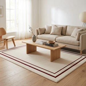 Puede incluir: Alfombra rectangular color crema con borde rojo oscuro en un salón. La alfombra está colocada debajo de una mesa de centro de madera clara y un sofá beige con cojines a juego. La habitación tiene pisos de madera y una paleta de colores neutros.
